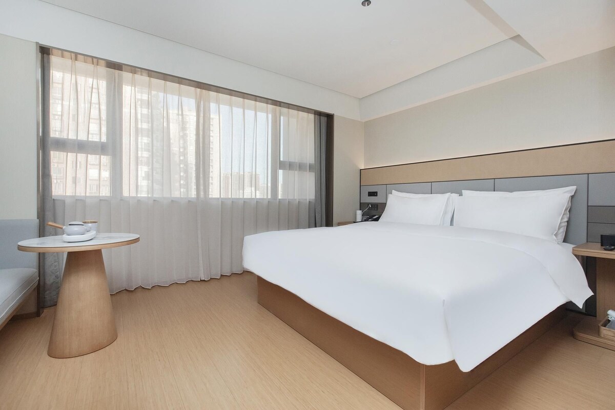 A modern hotel room with a large bed, a bedside table, a small round table, and a window with sheer curtains.