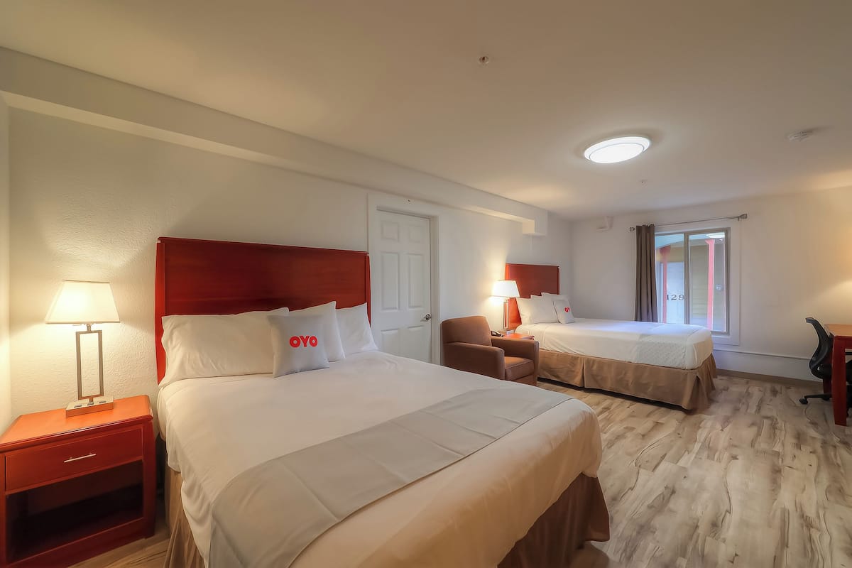 A hotel room with two beds, a nightstand with a lamp, a chair, and a desk.