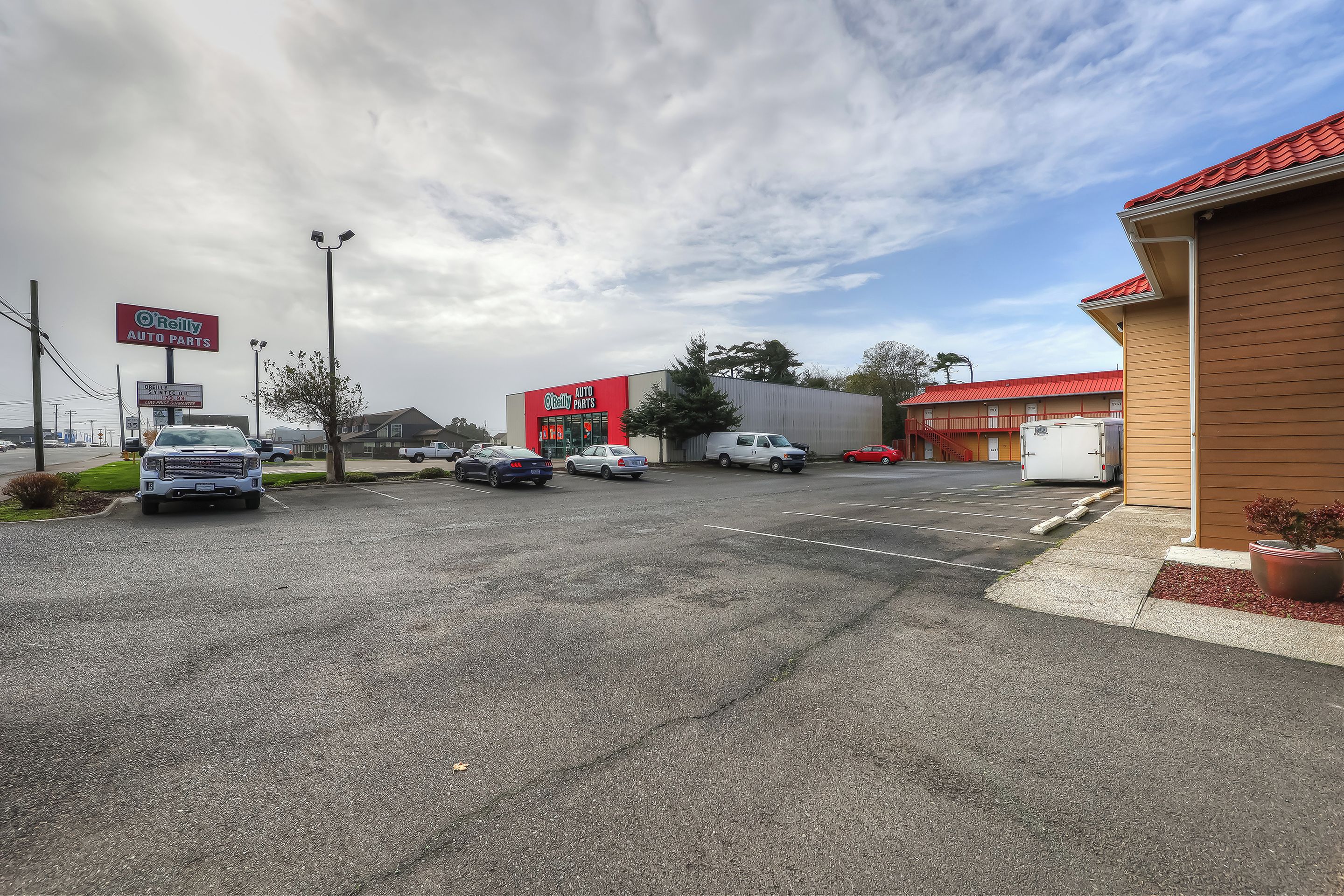 Un estacionamiento al aire libre con varios coches, un edificio con techo rojo y un cartel de 'O'Reilly Auto Parts'.