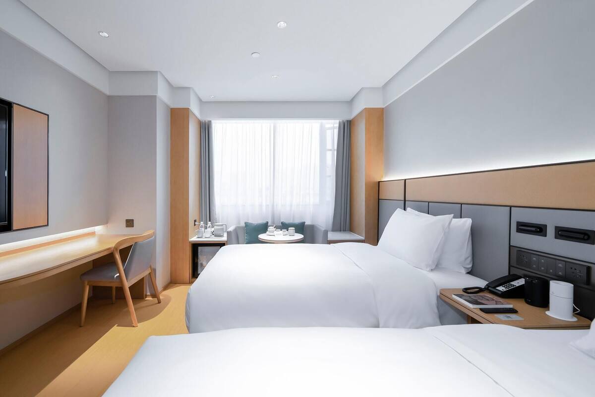 Hotellihuone, jossa on kaksi sänkyä, työpöytä, tuoli, pieni pöytä lampun kanssa ja suuri ikkuna, jossa on verhot.