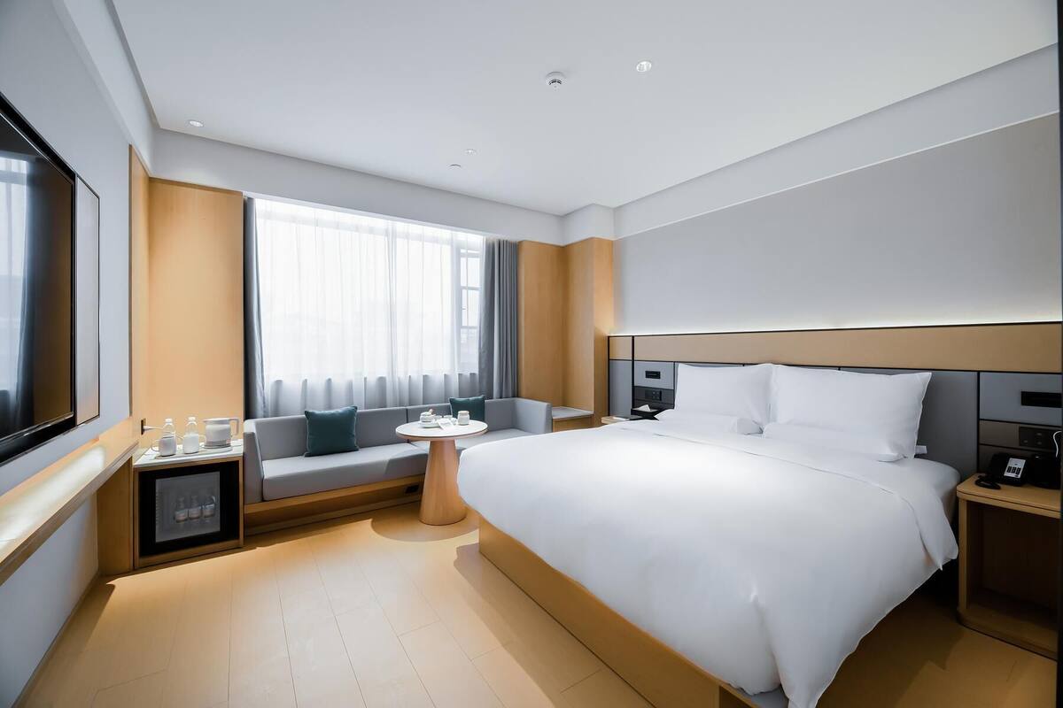 Moderni hotellihuone, jossa on suuri sänky, yöpöytä, pieni pöytä tuolilla, sohva ja suuri ikkuna, jossa on verhot.