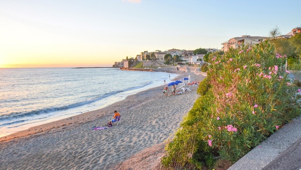Une plage avec une personne assise sur une chaise, une rangée de parasols et des bâtiments en arrière-plan.