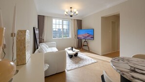 Ein modernes Wohnzimmer mit einem weißen Sofa, einem Fernseher auf einem Schrank, einem Esstisch und Blick auf die Stadt durch das Fenster.