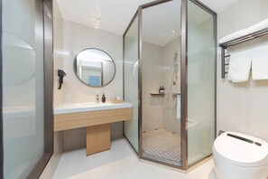 Un baño moderno con ducha envuelta en vidrio, un espejo redondo, un lavabo con tocador de madera y un inodoro.
