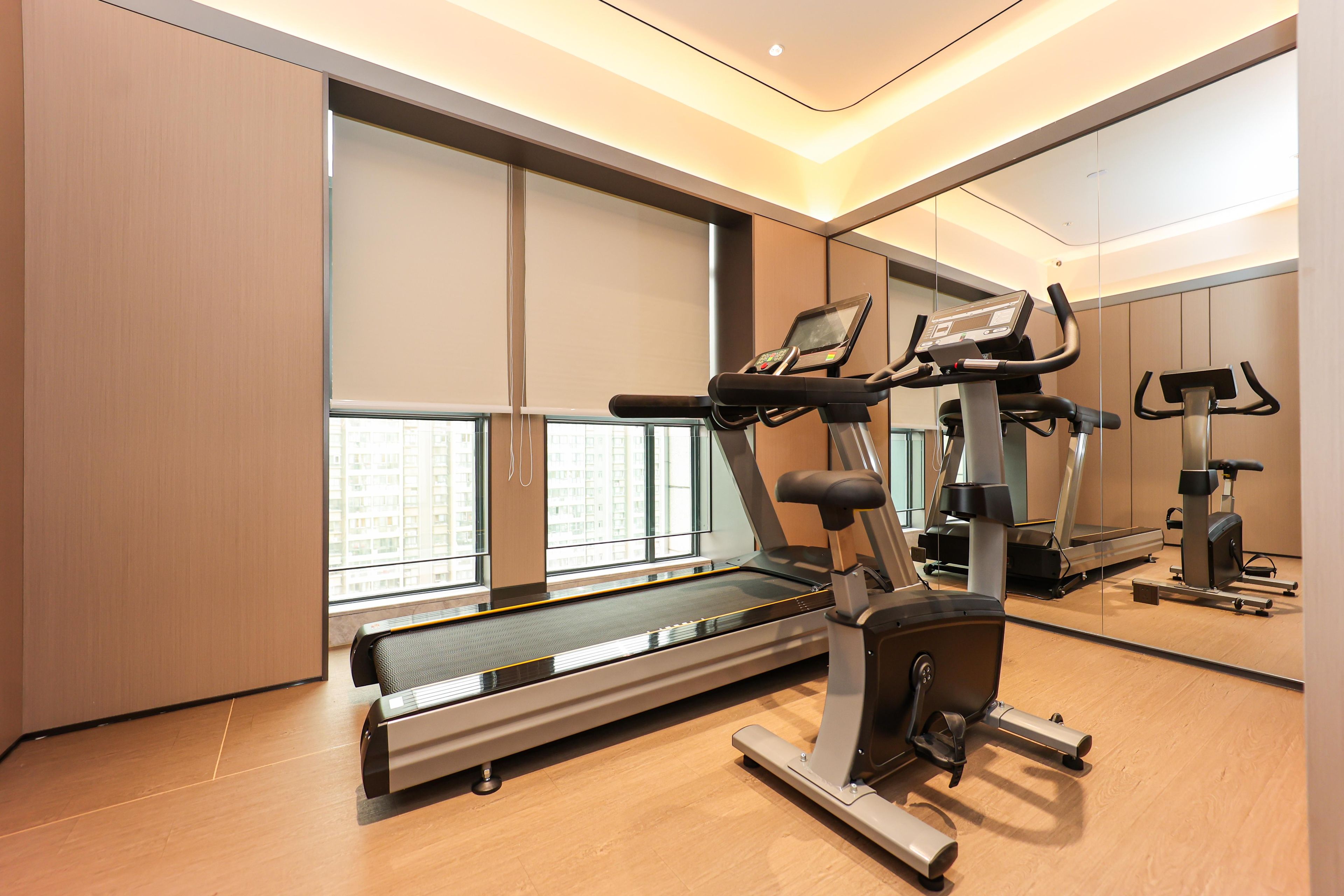 A modern gym with a treadmill, elliptical, and stationary bike, large windows, and a wooden floor.