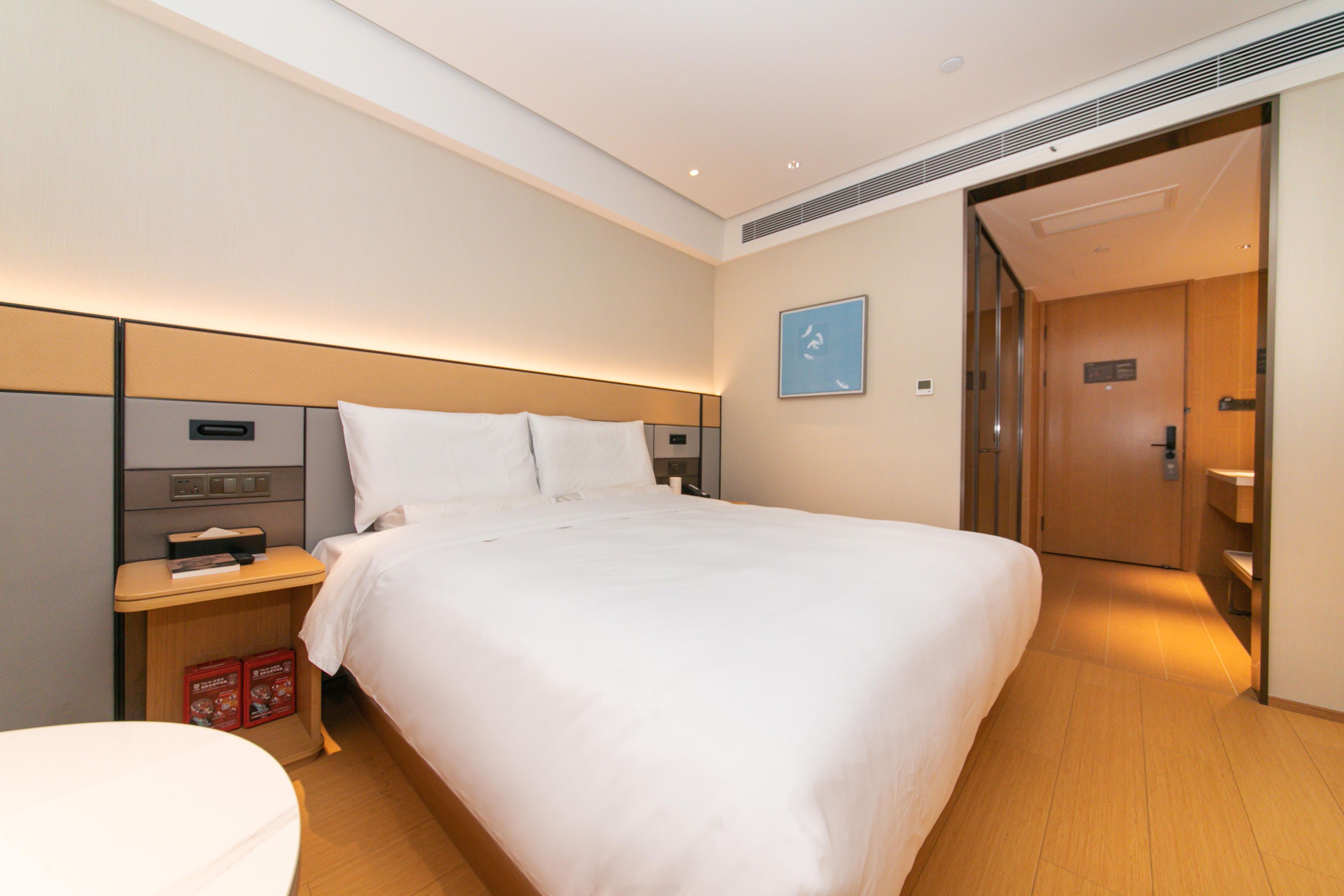 A hotel room with a large bed, a bedside table with a lamp, a small desk, and a trash bin.
