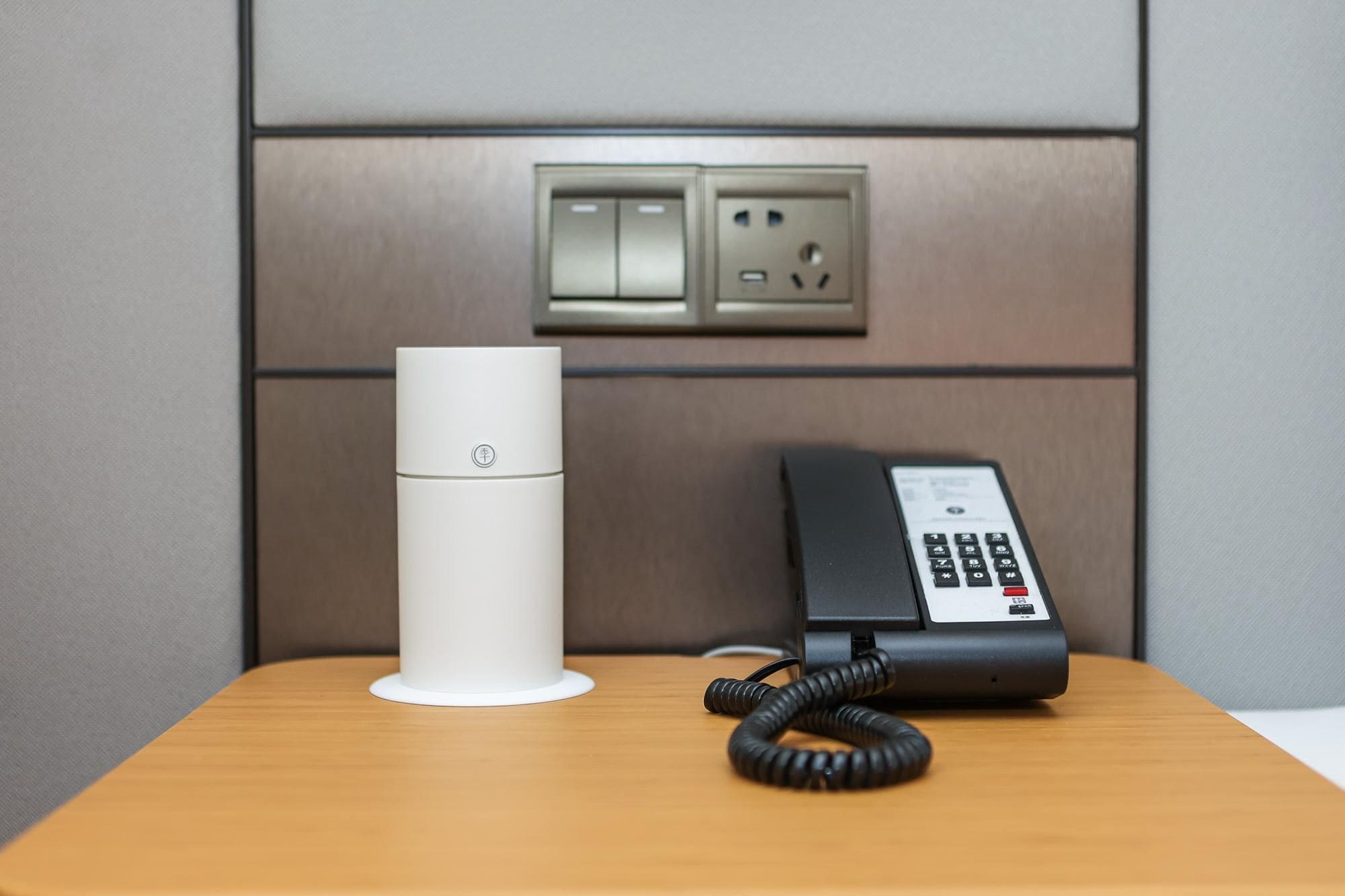 A white cylindrical device and a black telephone on a wooden surface, with a wall-mounted electrical outlet above them.