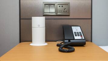 A white cylindrical device and a black telephone on a wooden surface, with a wall-mounted electrical outlet above them.