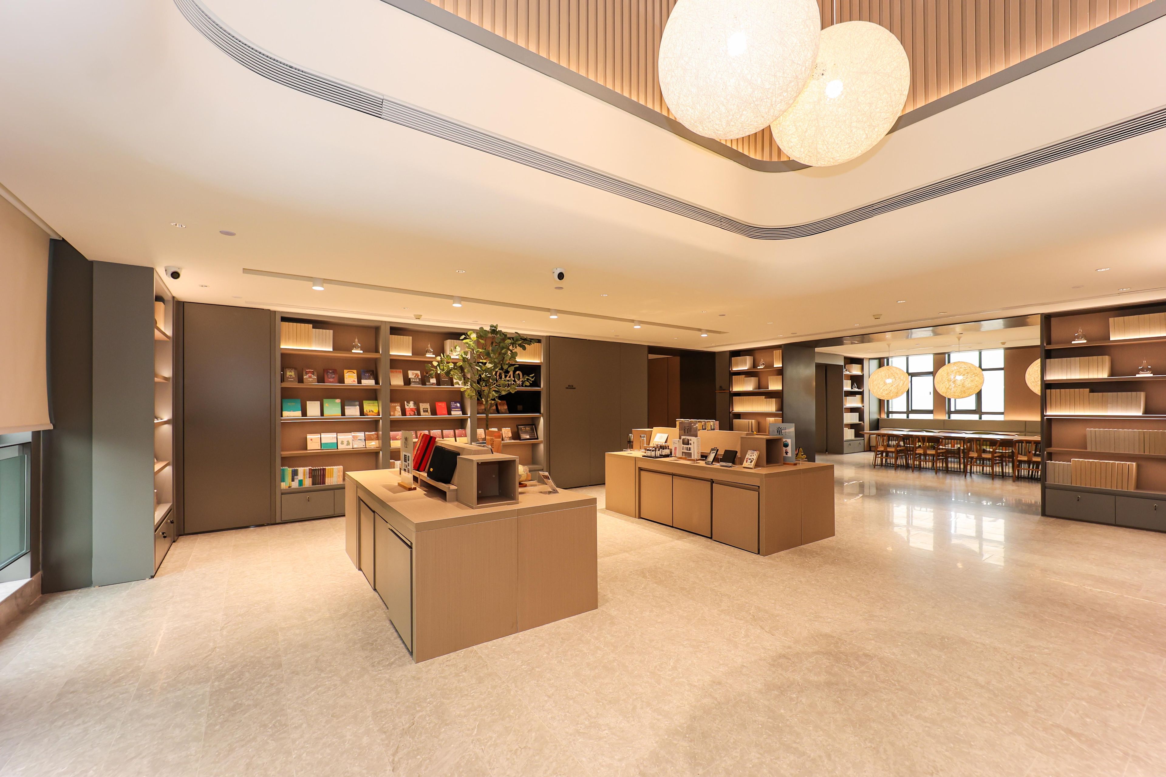 A modern hotel lobby with a central reception desk, shelves with books and decorative items, and a seating area with tables and chairs.