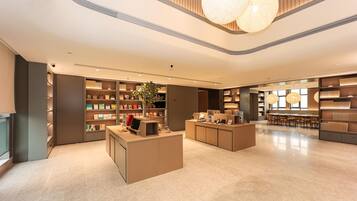 A modern hotel lobby with a central reception desk, shelves with books and decorative items, and a seating area with tables and chairs.