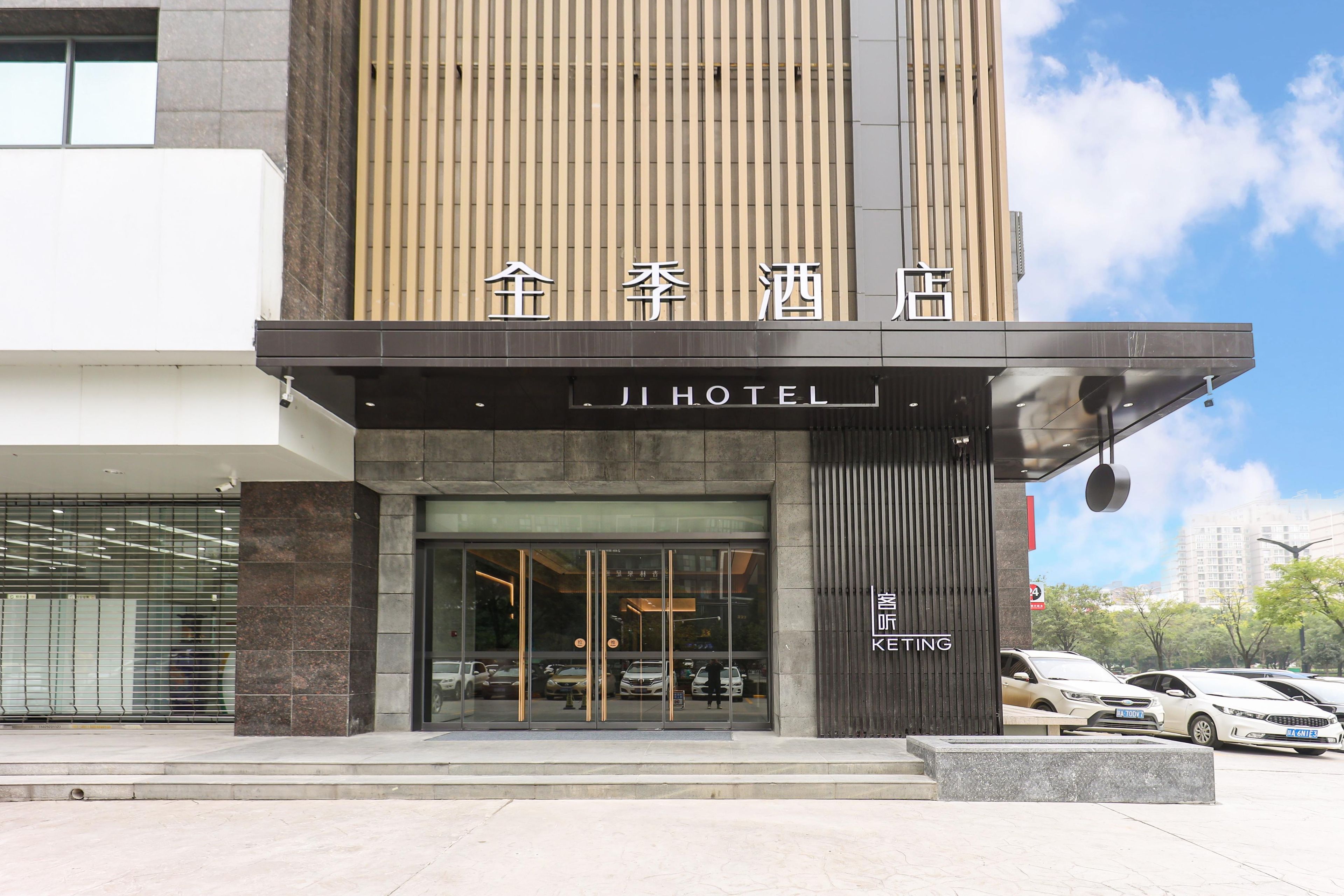 A hotel entrance with a modern design, featuring a large glass facade, a prominent sign, and a parking area with several cars.