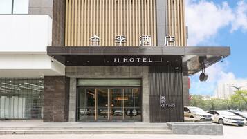 A hotel entrance with a modern design, featuring a large glass facade, a prominent sign, and a parking area with several cars.