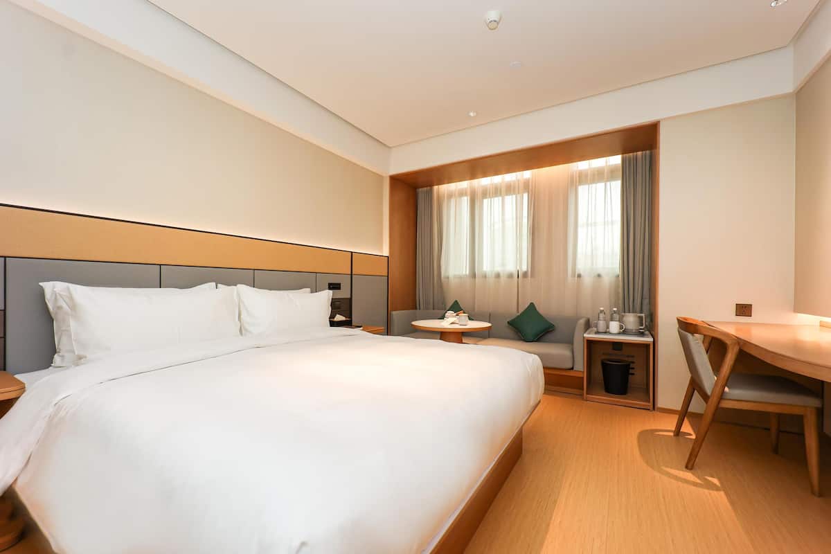 Habitación de hotel con una cama grande, un escritorio, una silla, una mesita, una ventana con cortinas y un cesto de basura.