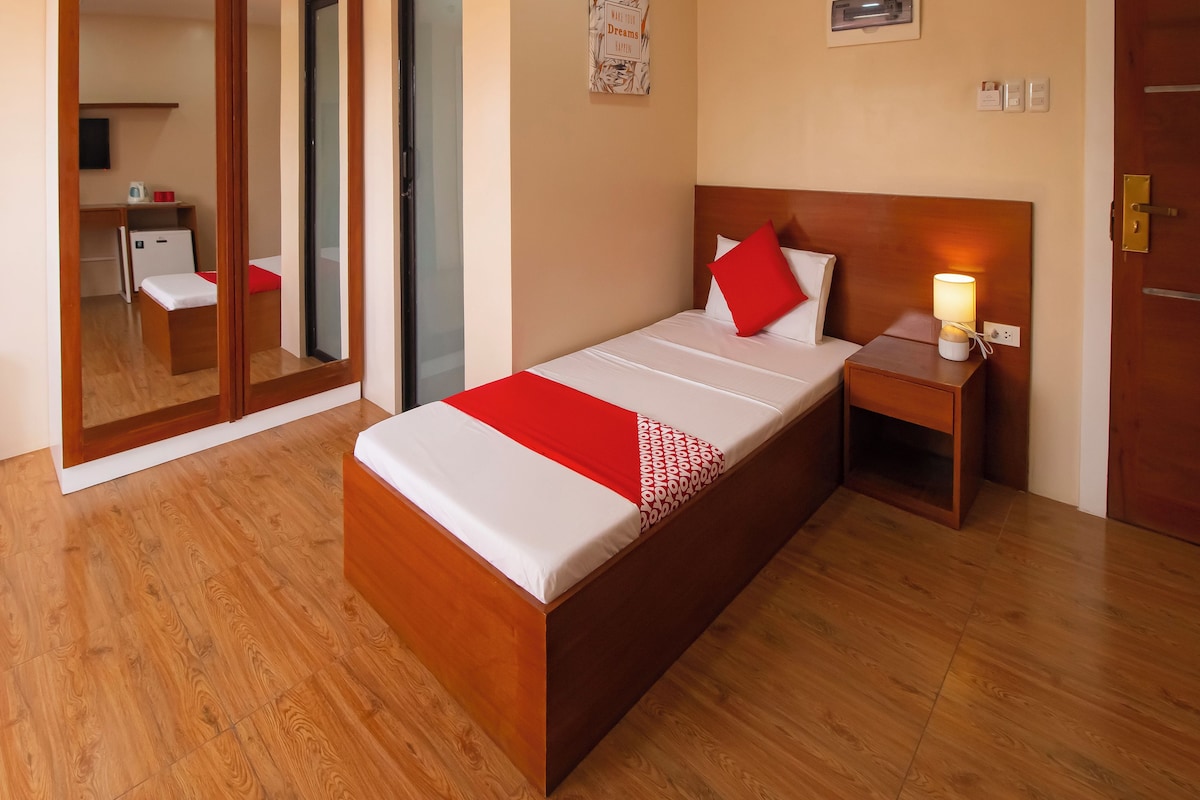 A hotel room with a wooden bed, a bedside table with a lamp, a mirror, and a small kitchenette in the background.