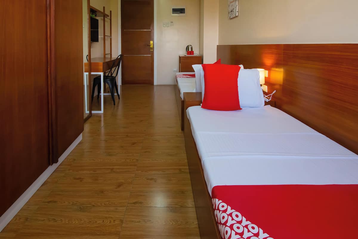 Habitación de hotel con piso de madera, una cama con almohada roja y blanca, una mesita de noche con lámpara y un armario de madera.