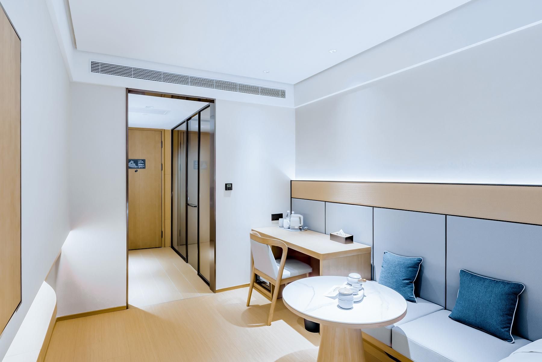 Ein modernes Hotelzimmer mit einem Holztisch, einem runden Tisch, einer Couch mit blauen Kissen und einer Schiebetür zu einem weiteren Raum.