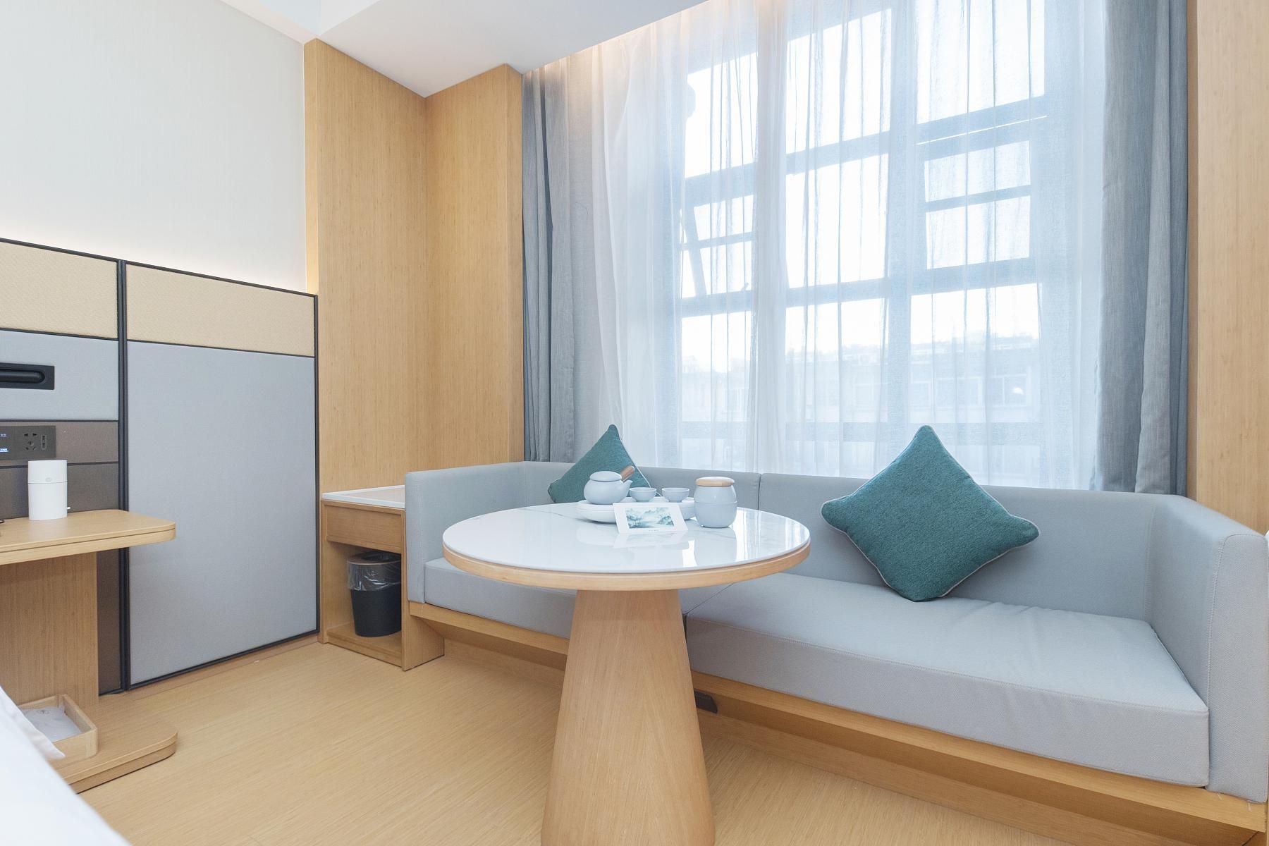 Una habitación de hotel moderna con kitchenette, una mesa de comedor redonda, un sofá con cojines y un gran ventanal con cortinas.