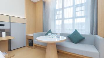 Una habitación de hotel moderna con kitchenette, una mesa de comedor redonda, un sofá con cojines y un gran ventanal con cortinas.