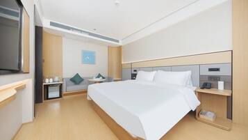 Habitación de hotel con una cama grande, una mesita de noche con teléfono y lámpara, un pequeño rincón de descanso con sofá y mesa, y un televisor montado en la pared.