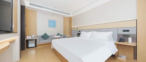 Quarto de hotel com uma cama grande, uma mesa de cabeceira com telefone e luminária, um pequeno espaço de estar com sofá e mesa, e uma televisão na parede.