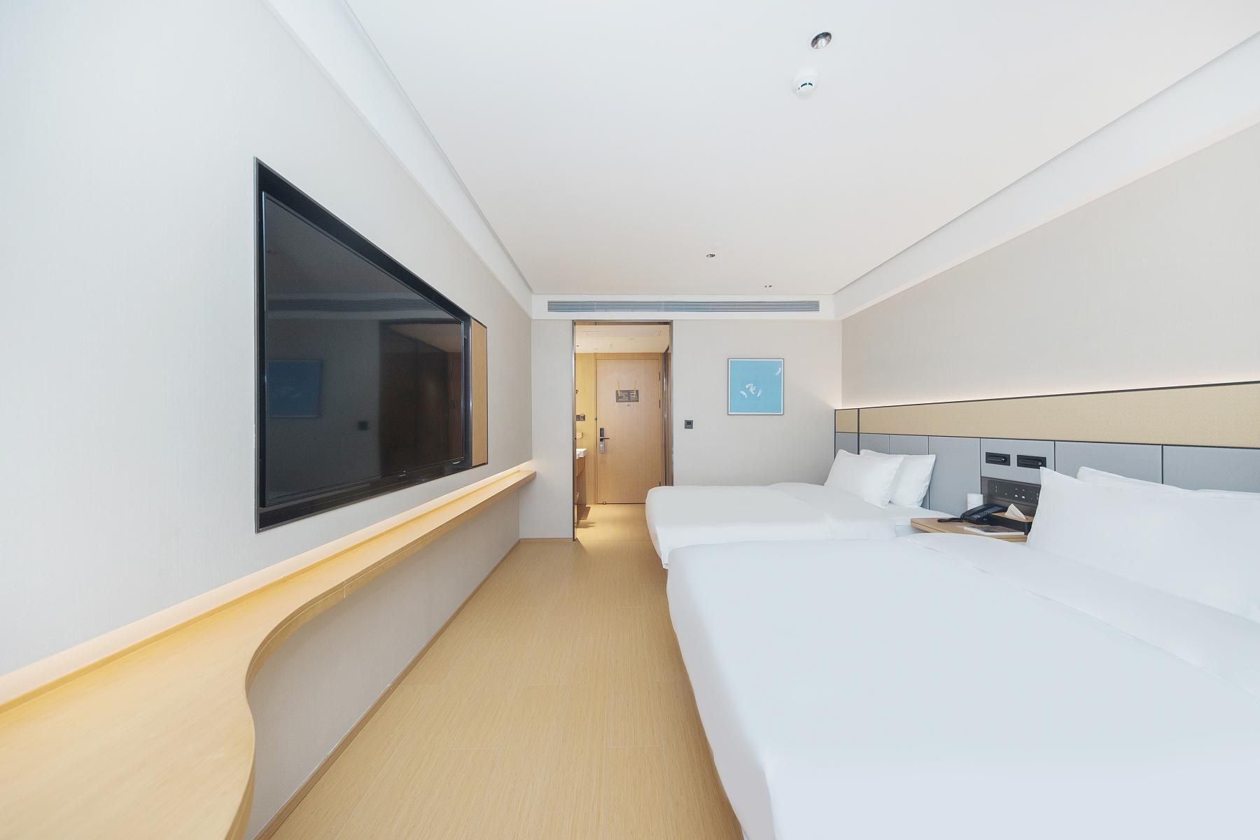 A hotel room with a large window, a bed, a bedside table, and a door leading to the hallway.