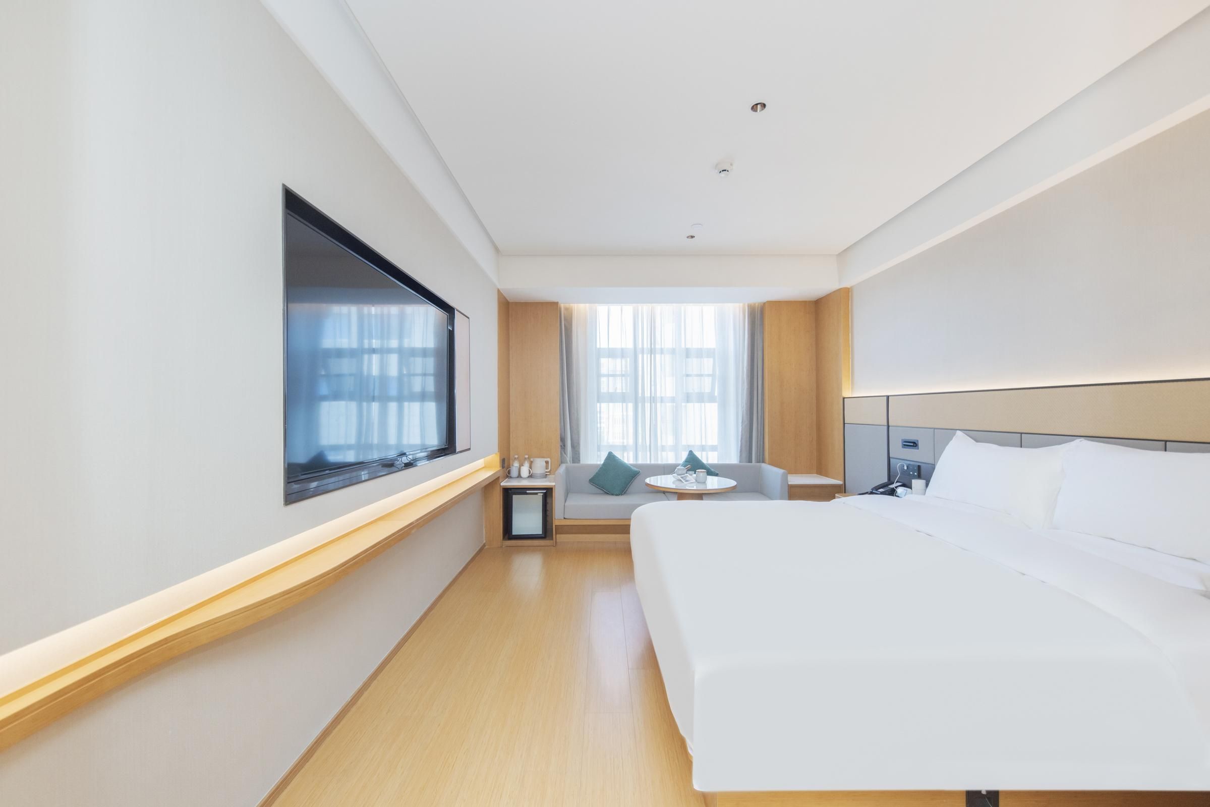 Habitación de hotel con una cama grande, un televisor montado en la pared, una mesita con una silla y una ventana con cortinas.