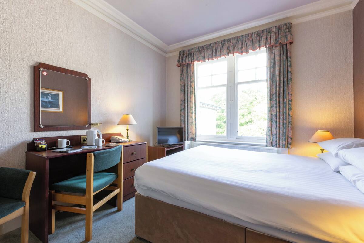 Ett hotellrum med en säng, ett skrivbord, en stol, en tv och ett fönster med gardiner.