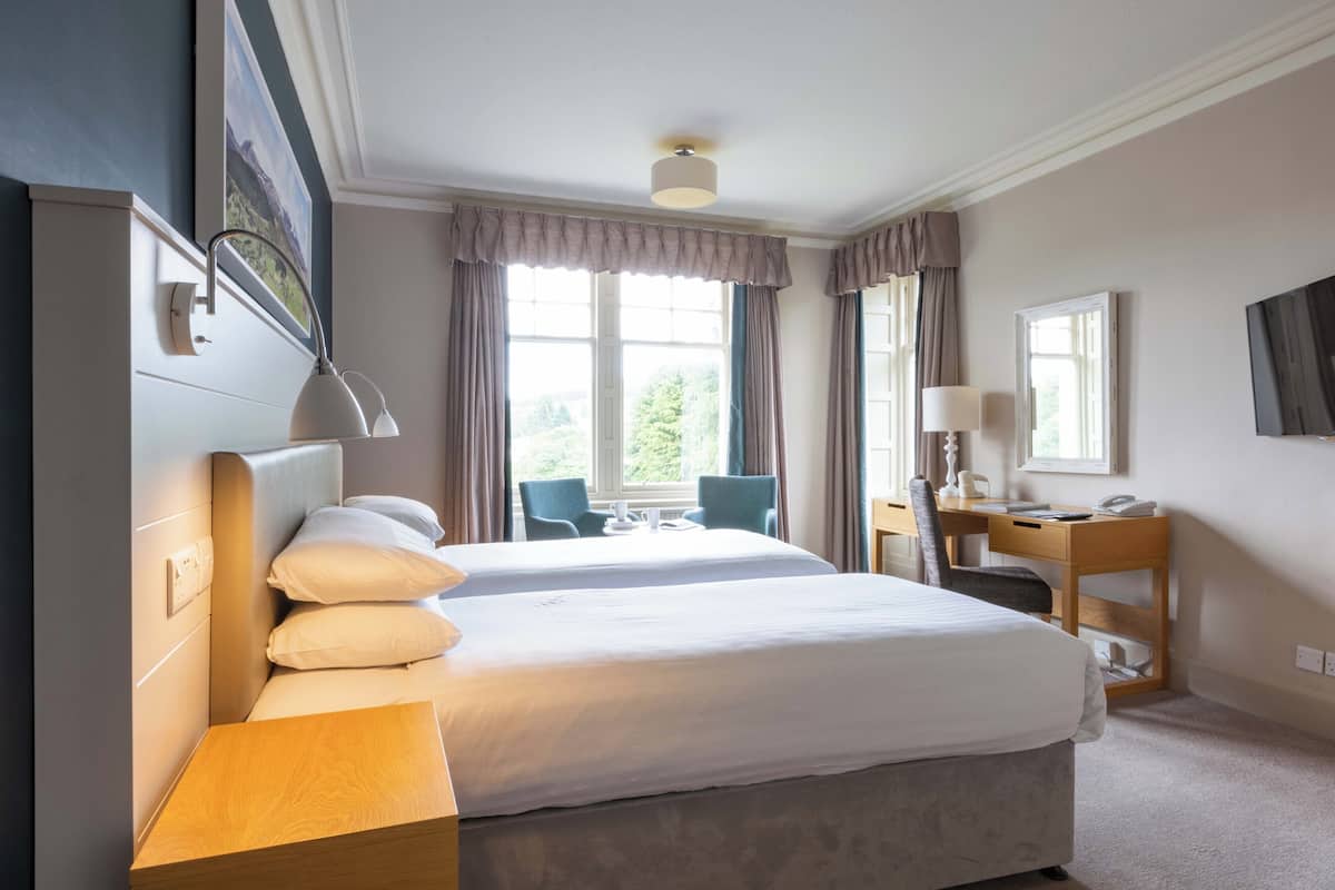 Ett hotellrum med två sängar, ett skrivbord, en stol, en TV och ett fönster med gardiner.