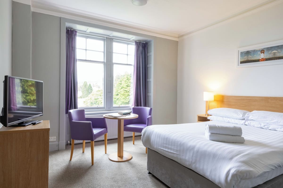 Ett hotellrum med en säng, en tv, ett skrivbord, två stolar, ett bord och ett fönster med gardiner.
