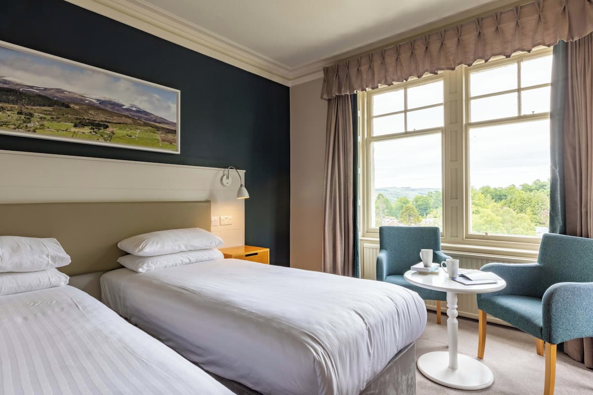 Ett hotellrum med två sängar, ett litet bord med en stol, en tavla på väggen och ett fönster med gardiner.