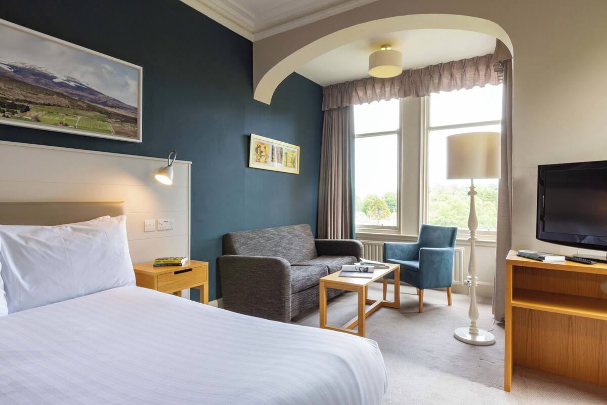 Ett hotellrum med en säng, en soffa, ett skrivbord, en tv och ett fönster med gardiner.