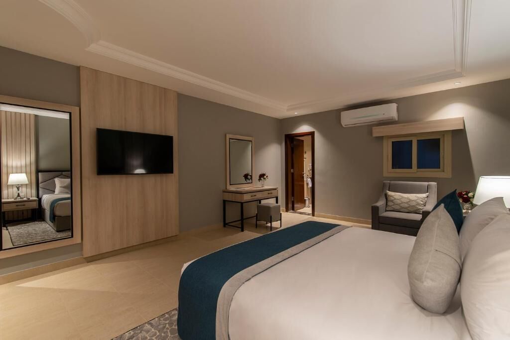 A hotel room with a large bed, a desk with a chair, a television, a mirror, and an air conditioning unit.