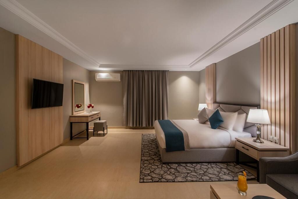 A hotel room with a bed, bedside table, lamp, TV, and a desk with a chair.