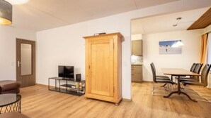Ein Wohnzimmer mit Fernseher, Schrank, Essbereich mit Stühlen und einer Küche im Hintergrund.