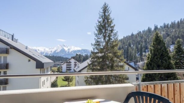 Ein Balkon mit gedecktem Tisch, mit Blick auf eine gebirgige Landschaft mit Gebäuden und Bäumen.