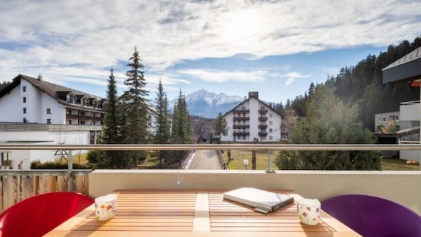 A balcony with a wooden table and two chairs, overlooking a mountain landscape with buildings and trees. - Val Signina 5-2 by Interhome (Laax)