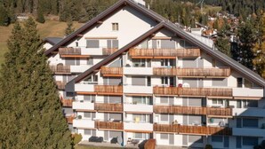 Ein mehrgeschossiges Gebäude mit Balkonen, umgeben von Bäumen und einem Bergpanorama.