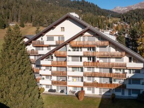 Ein mehrgeschossiges Gebäude mit Balkonen, umgeben von Bäumen und einem Bergpanorama.