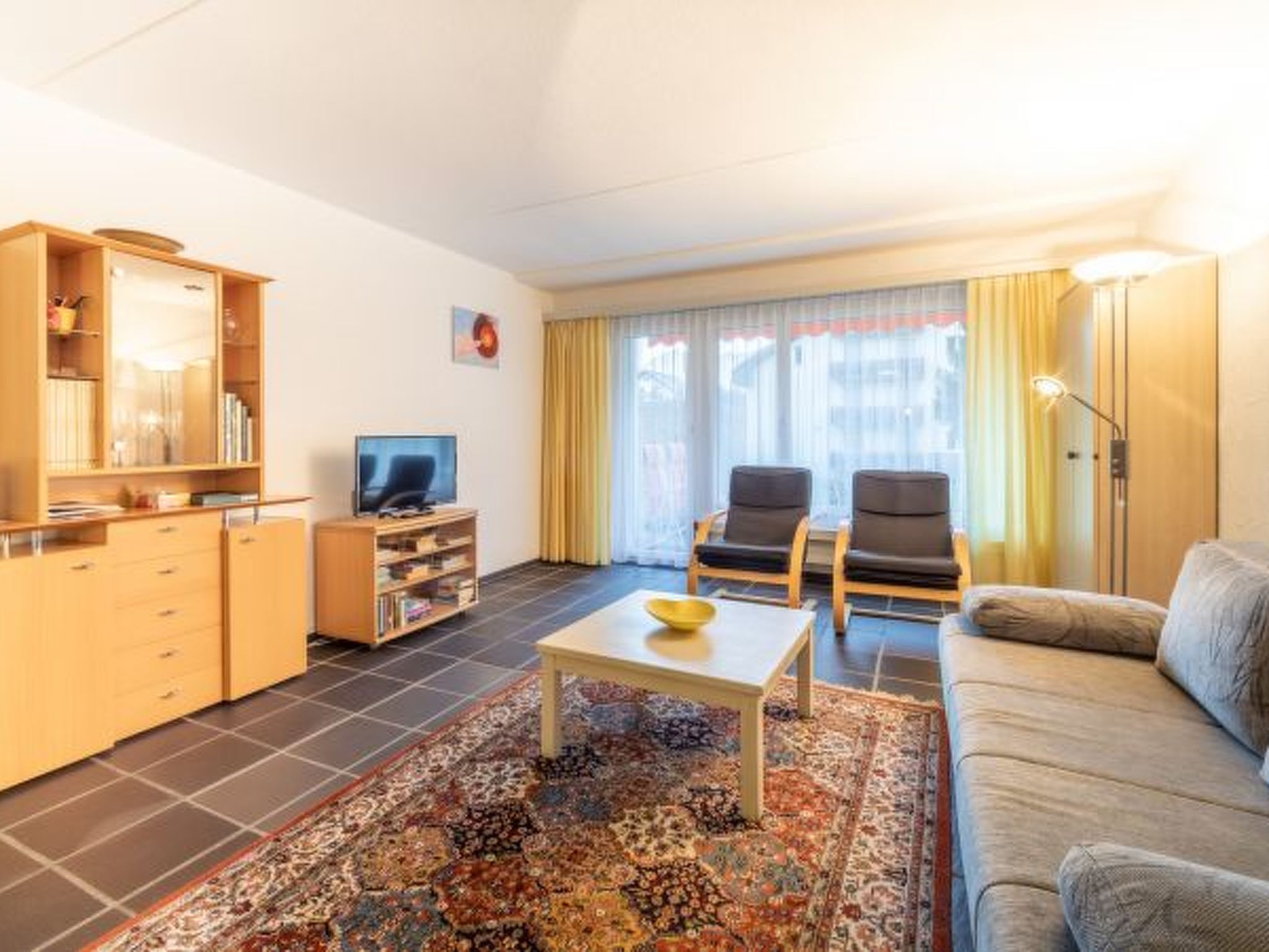 Una sala de estar con un sofá, dos sillones, una mesa de centro, un televisor, una estantería y una alfombra estampada.