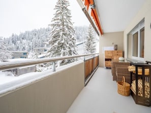 A balcony with a view of snow-covered trees and buildings, a wooden chair, a wicker basket, and a small table with a lamp. - Val Signina 7-12 by Interhome (Laax)