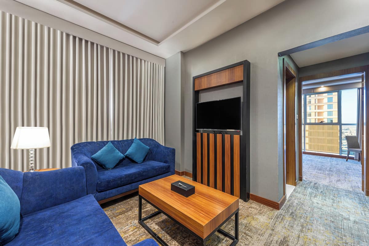 Une chambre d’hôtel moderne avec un canapé bleu, une table basse en bois, un téléviseur à écran plat et une vue sur le balcon extérieur.