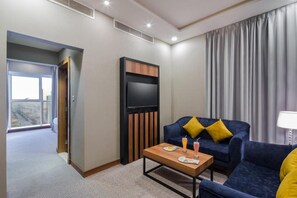 Une chambre d’hôtel équipée d’un téléviseur à écran plat, d’un canapé avec des coussins jaunes, d’une table basse avec des boissons et d’une grande fenêtre avec des rideaux.