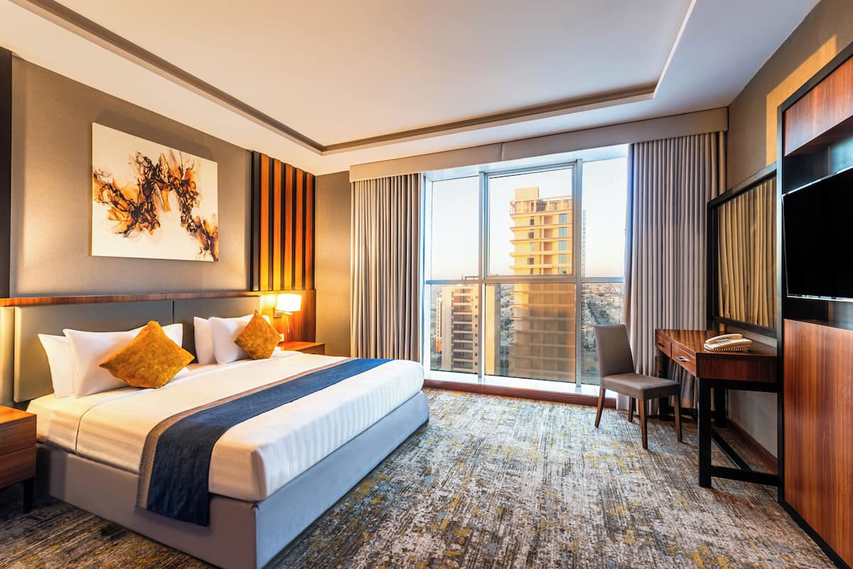 Une chambre d’hôtel avec un grand lit, un bureau, une chaise, une télévision et une vue sur les bâtiments par la fenêtre.