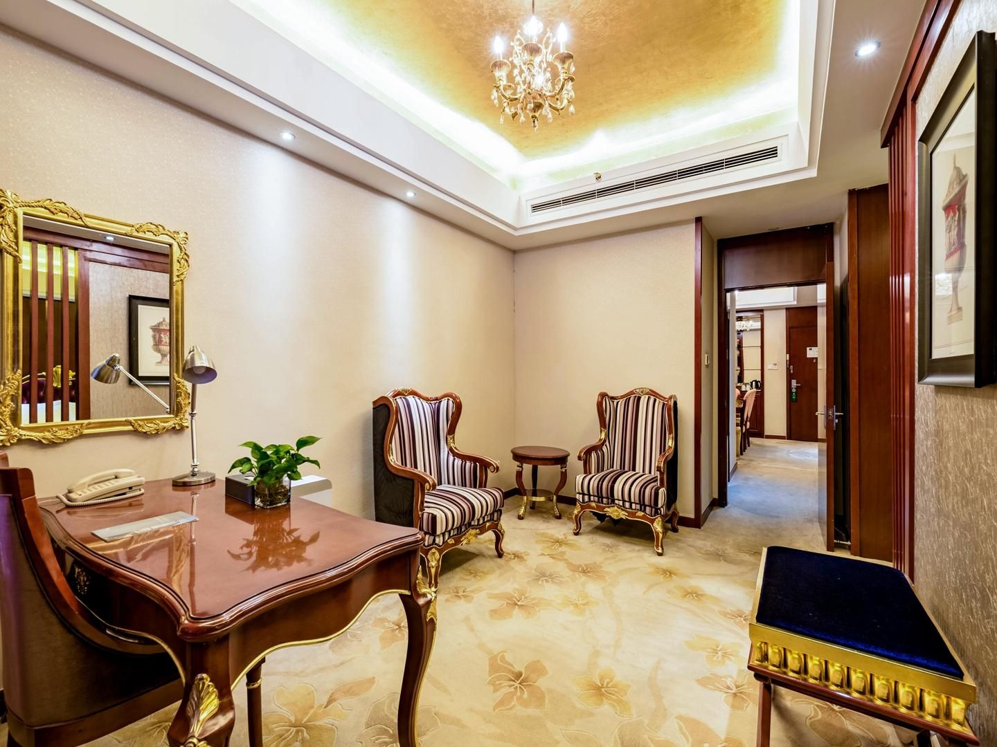 Un vestíbulo de hotel con un mostrador de recepción de madera, dos sillones tapizados, una mesita redonda y un espejo grande con marco dorado.
