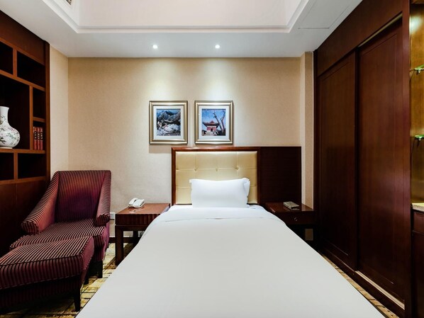 Quarto de hotel com cama, uma cadeira, mesa de cabeceira, telefone e duas imagens emolduradas na parede.