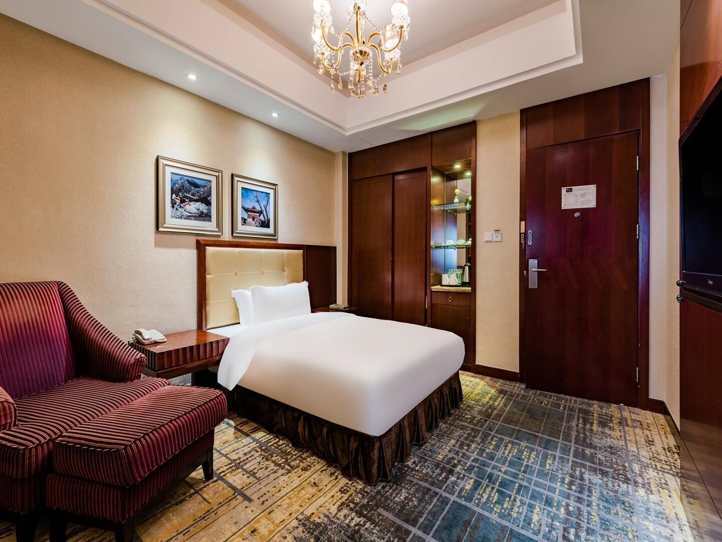 A hotel room with a large bed, a red upholstered chair, a wooden nightstand, a chandelier, and a wardrobe.