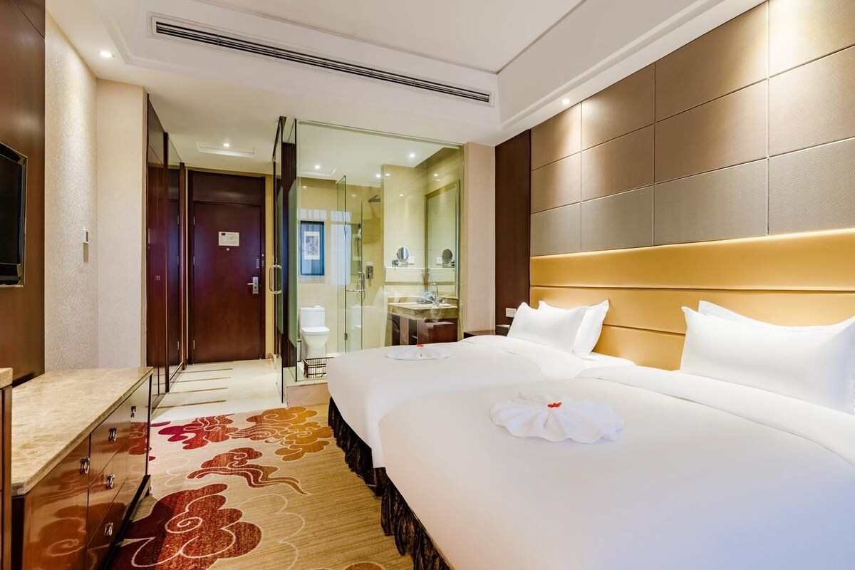 A hotel room with a large bed, a desk, a mirror, and a bathroom visible through a glass door.