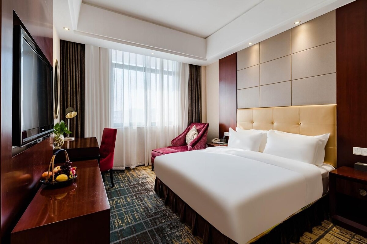 A hotel room with a large bed, a desk with a TV, a chair, and a window with curtains.