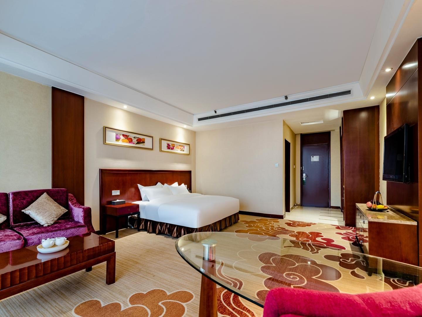 A hotel room with a bed, a sofa, a glass coffee table, a TV, and a door leading to another room.
