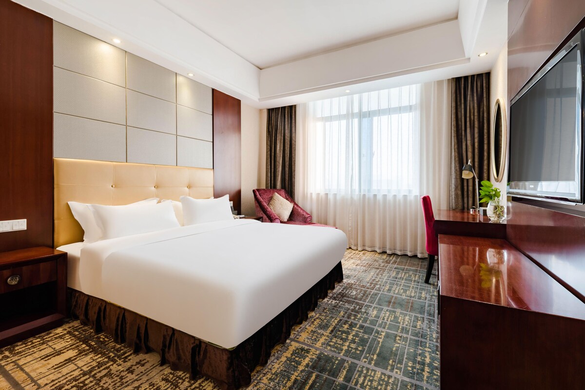 A hotel room with a large bed, a desk, a chair, a TV, and a window with curtains.